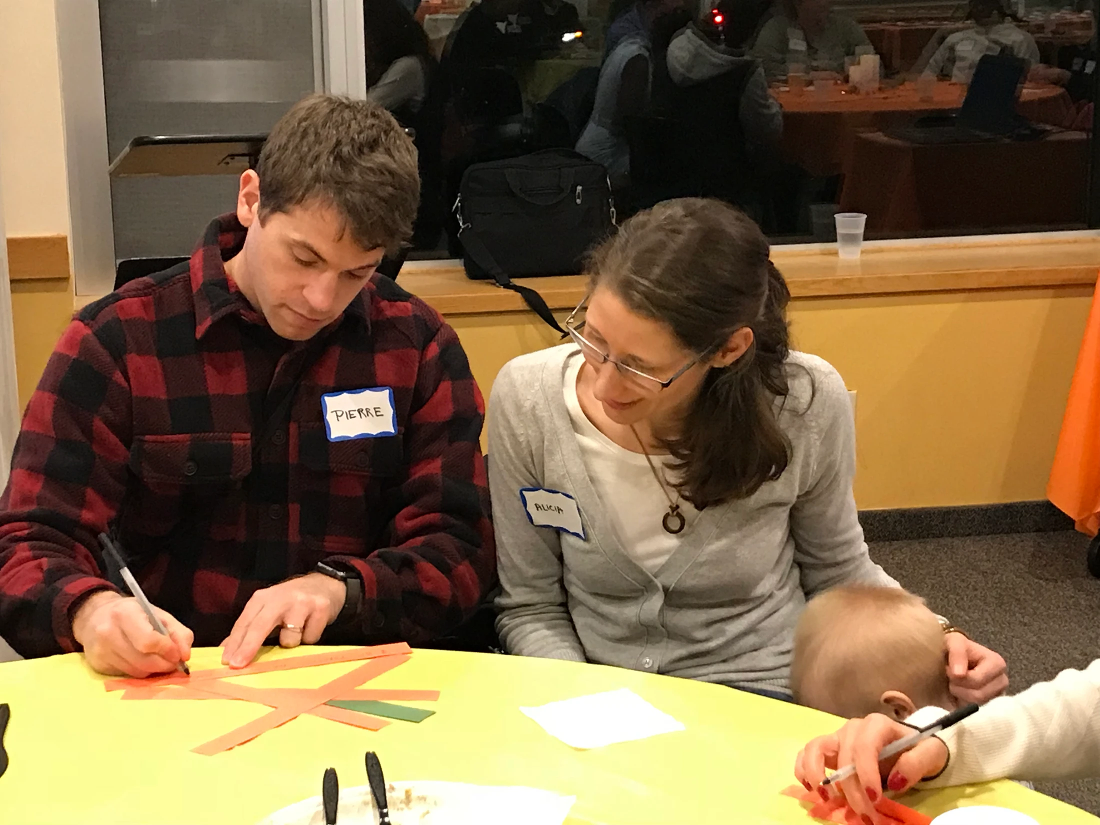 A man in a red plaid shirt and a woman in a grey cardigan sit at a yellow table doing a craft activity, with a small child also present.