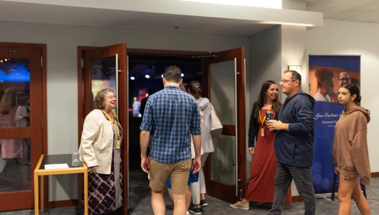 A group of people in casual attire talk and walk in a hallway near a set of double doors leading into a conference room.