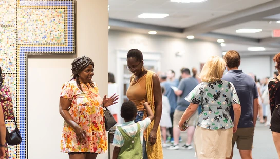 Two Black women and a child talk in a busy hallway near a mosaic cross. Other people are mingling in the background at an indoor event.