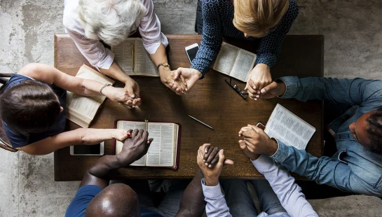 An overhead view of a diverse group of six people sitting around a wooden table, holding hands in prayer with open Bibles in front of them.