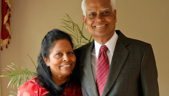 An older couple of South Asian descent smiling at the camera. The woman wears a red sari, and the man wears a dark suit with a red patterned tie.