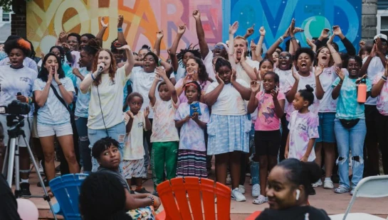 A large, diverse group of adults and children cheer with their hands in the air while standing in front of a colorful mural during an outdoor event.