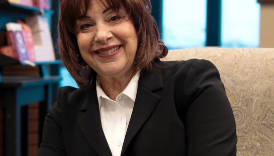 A woman with reddish-brown hair smiles warmly at the camera. She is wearing a black suit jacket and white collared shirt, and is seated in a patterned chair.