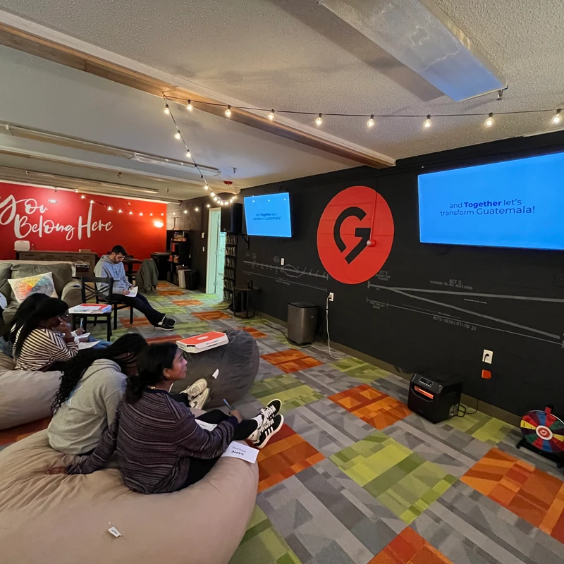 A diverse group of people sit on beanbags and a couch in a colorful room, watching a presentation about Guatemala on wall-mounted screens.