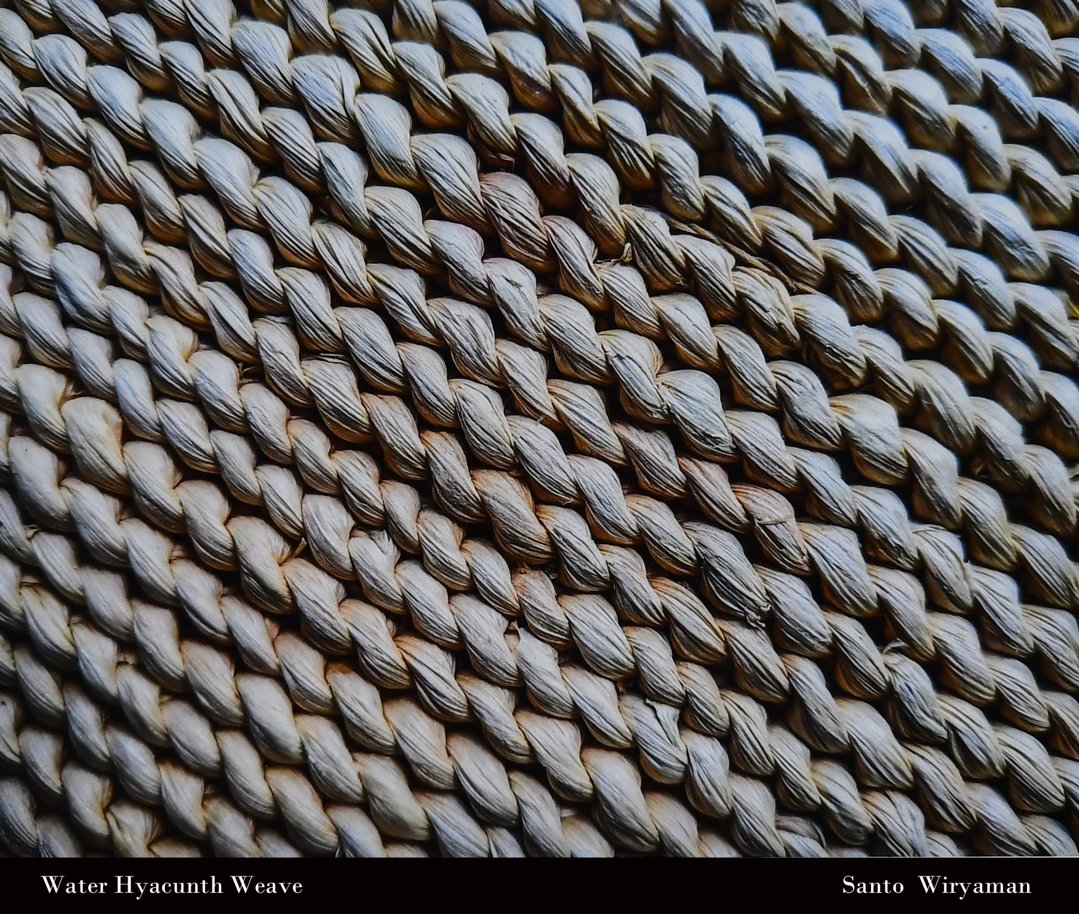 A detailed, close-up photograph of a tightly woven material made from dried water hyacinth, showing a diagonal pattern of braided fibers.
