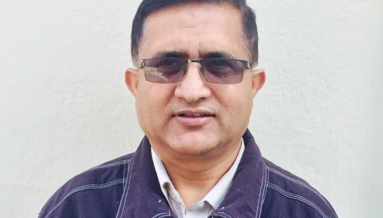 A head-and-shoulders portrait of a middle-aged man with short dark hair. He wears tinted glasses, a collared shirt, and a dark jacket against a plain wall.