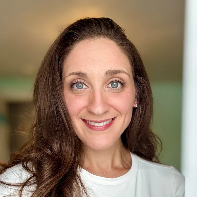 A close-up headshot of a woman with long, dark brown hair and bright blue eyes smiling directly at the camera. She is wearing a white t-shirt.