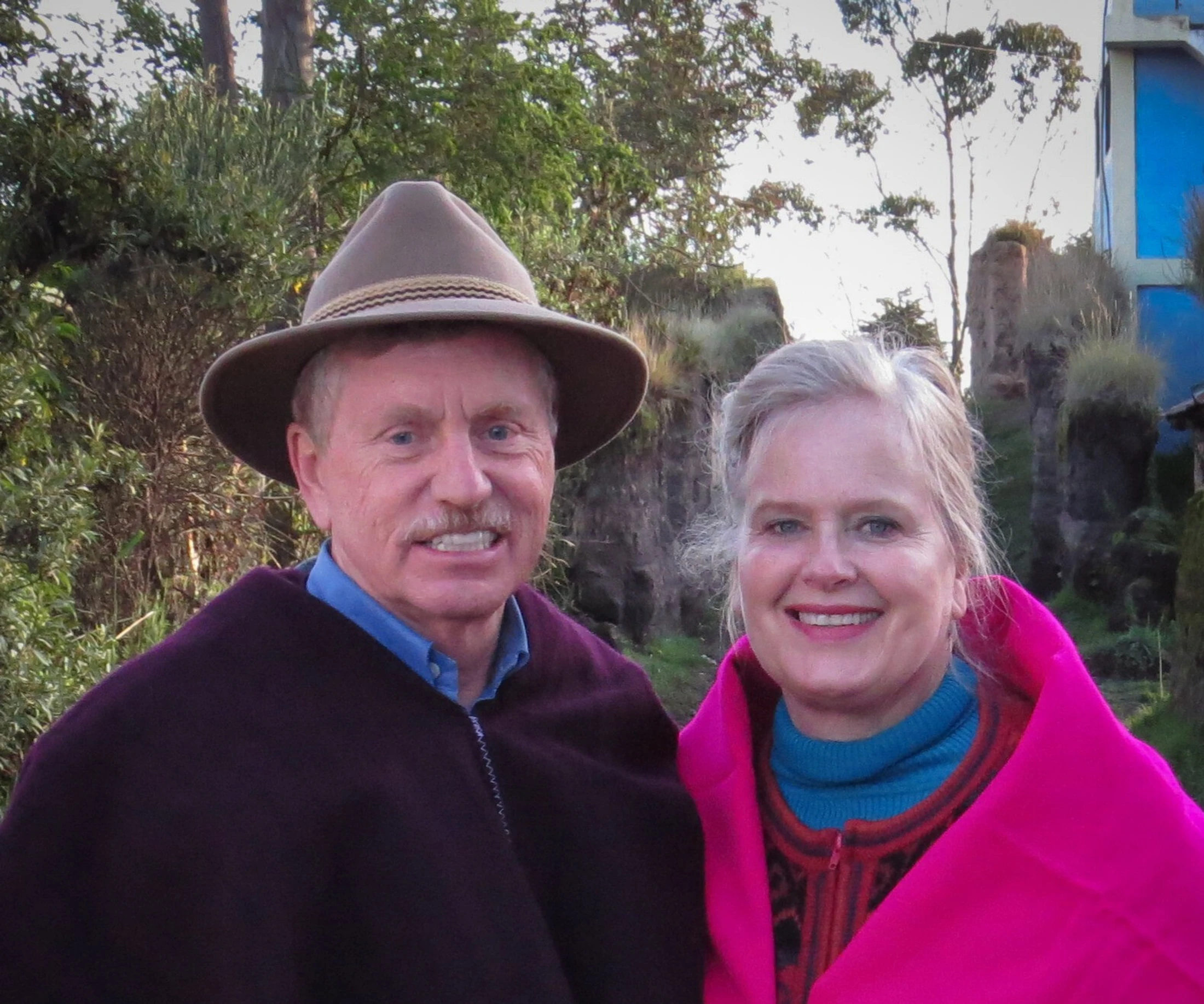 A smiling man and woman stand side-by-side outdoors. The man wears a brown hat and a purple poncho, and the woman is wrapped in a bright pink poncho.