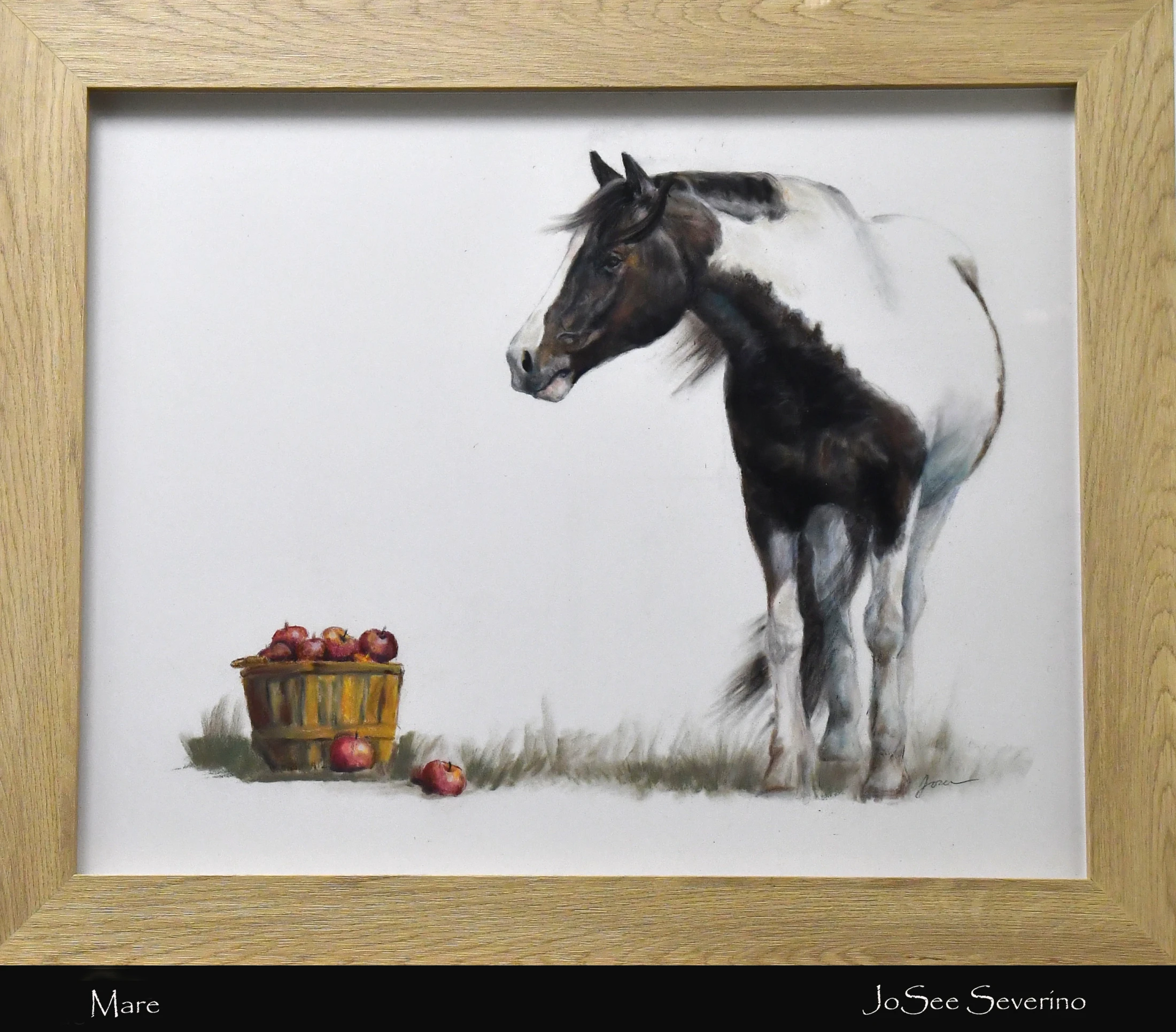 A framed painting by JoSee Severino of a black and white pinto mare standing in a grassy field next to a basket full of red apples.