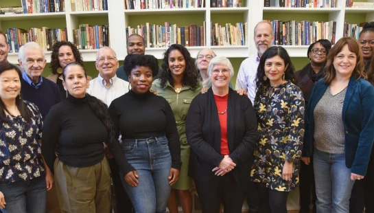 A diverse group of about fifteen people, men and women of various ages, stand together smiling for a photo in front of green and white bookshelves.