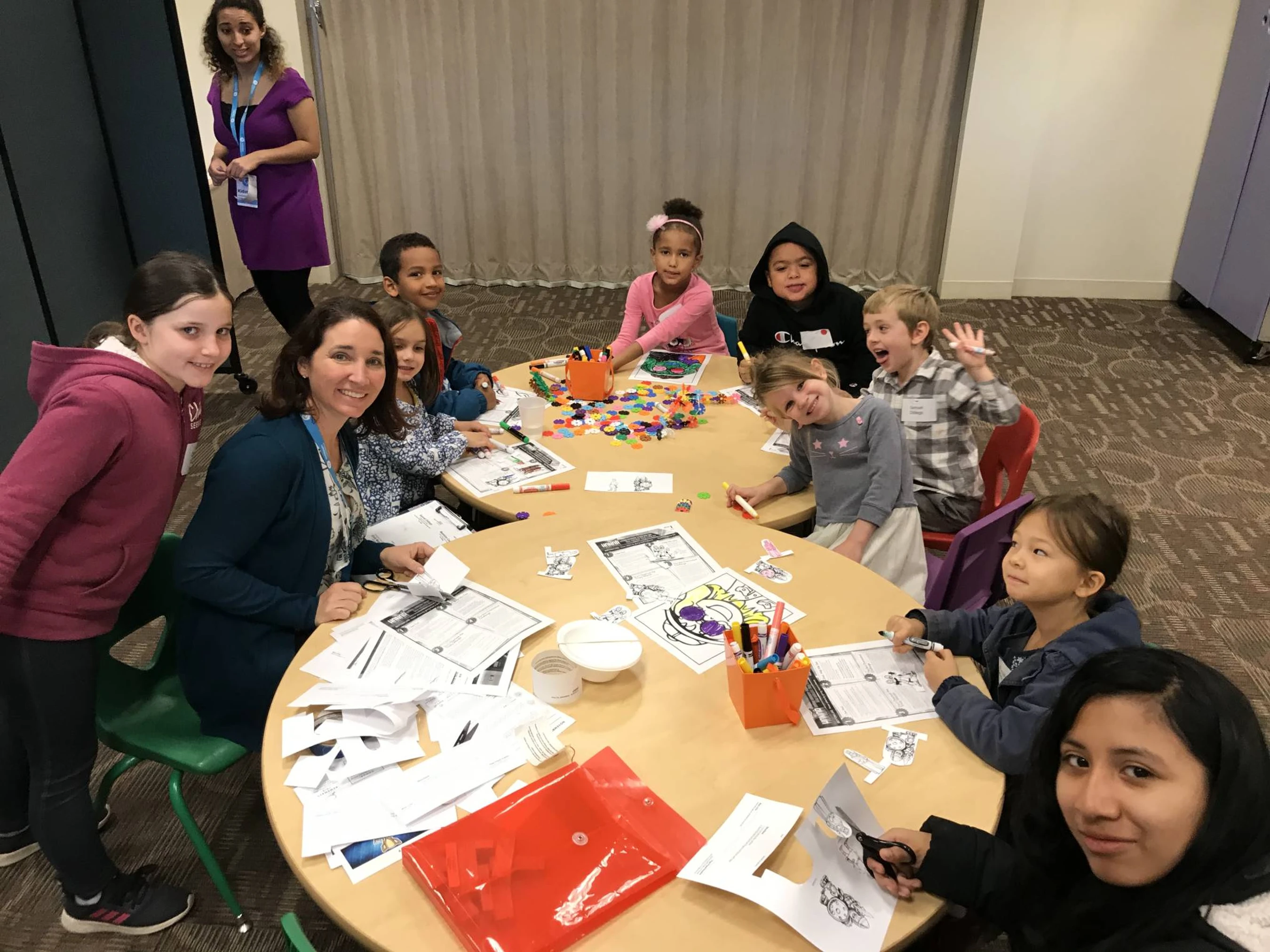 A diverse group of children and adults sit at round tables, smiling at the camera while working on coloring and cutting activities in a classroom setting.