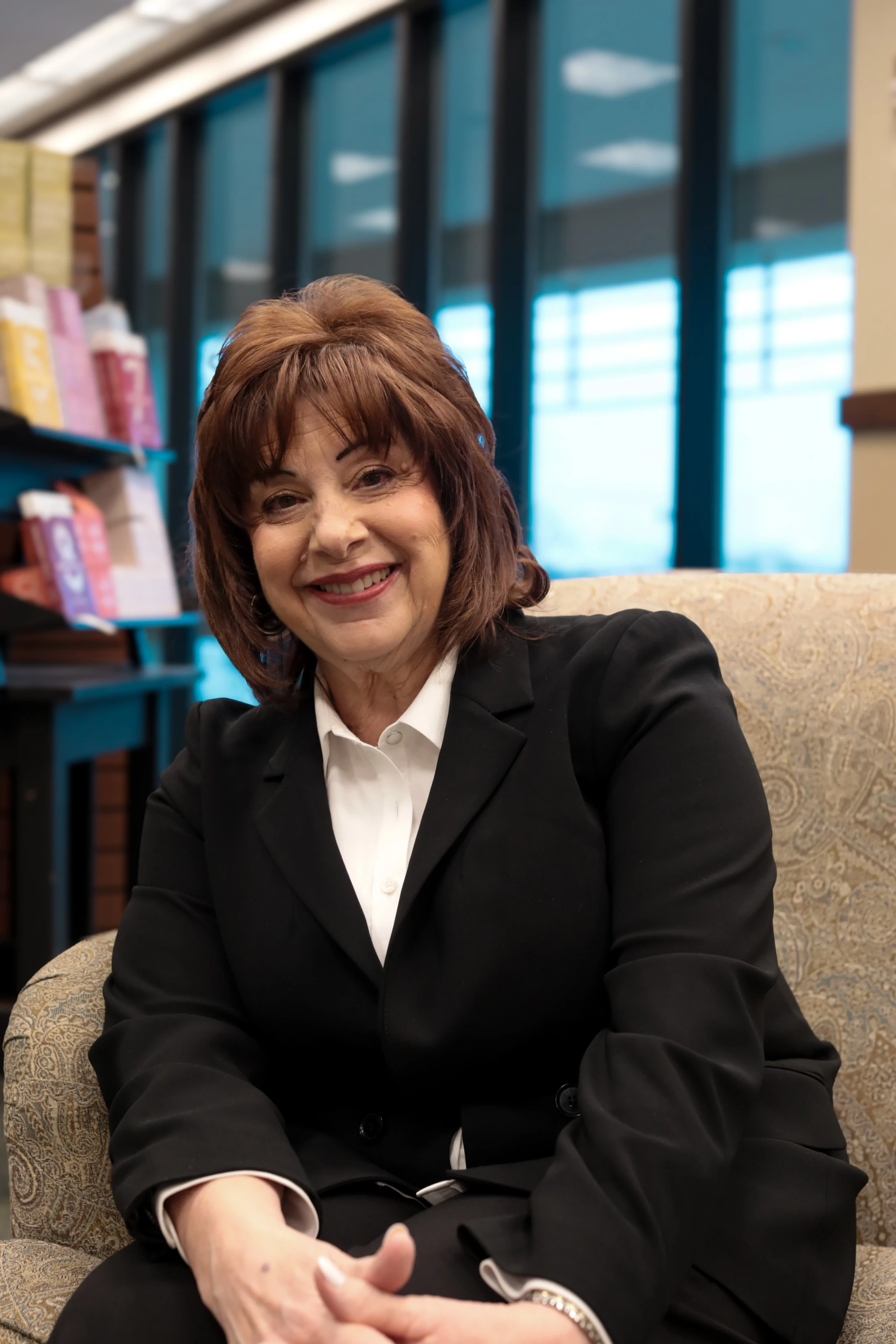 A woman with reddish-brown hair smiles warmly at the camera. She is wearing a black suit jacket and white collared shirt, and is seated in a patterned chair.