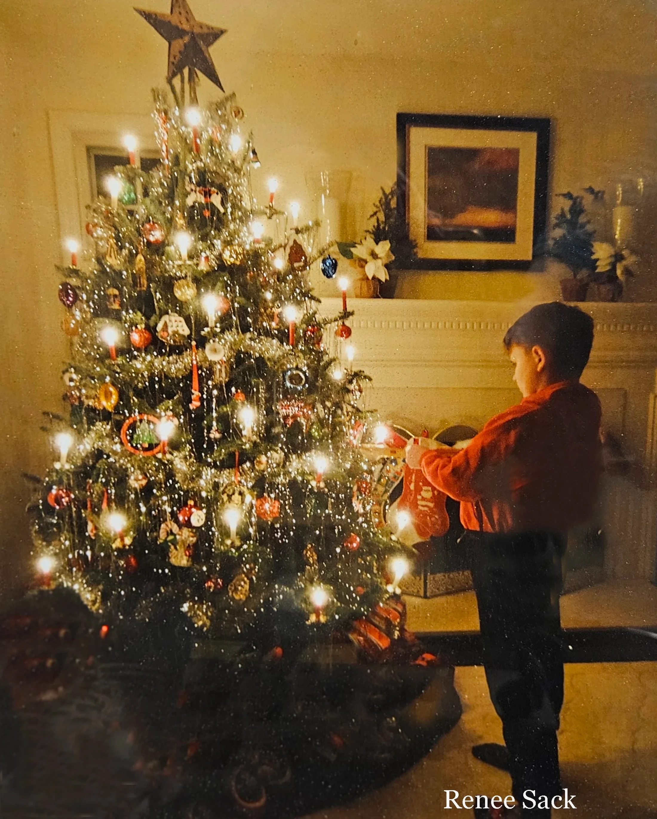 A vintage photograph shows a young boy in a red shirt standing by a brightly lit Christmas tree, holding a red stocking near a fireplace.