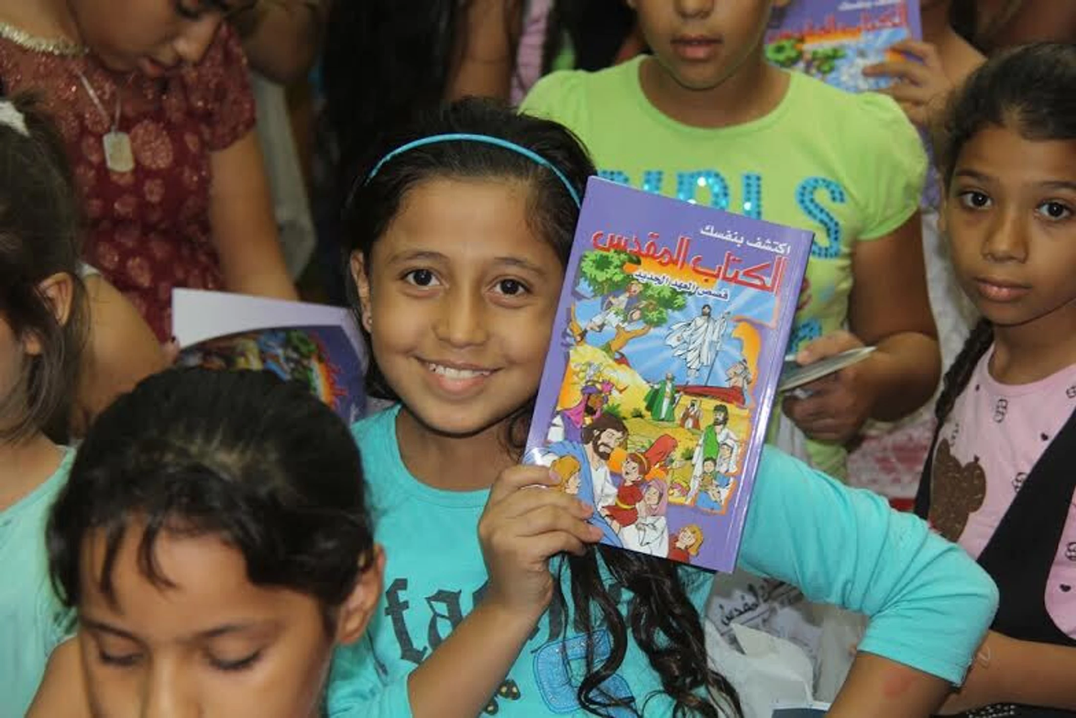 A smiling young girl in a blue shirt and headband holds up a colorful children's book with an Arabic title, surrounded by other children.
