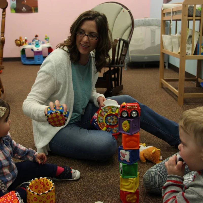 A woman sits on a carpeted floor with a young boy and girl. They are surrounded by colorful toys, including a tall stack of blocks, in a nursery setting.
