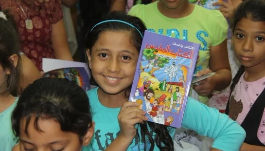 A smiling young girl in a blue shirt and headband holds up a colorful children's book with an Arabic title, surrounded by other children.