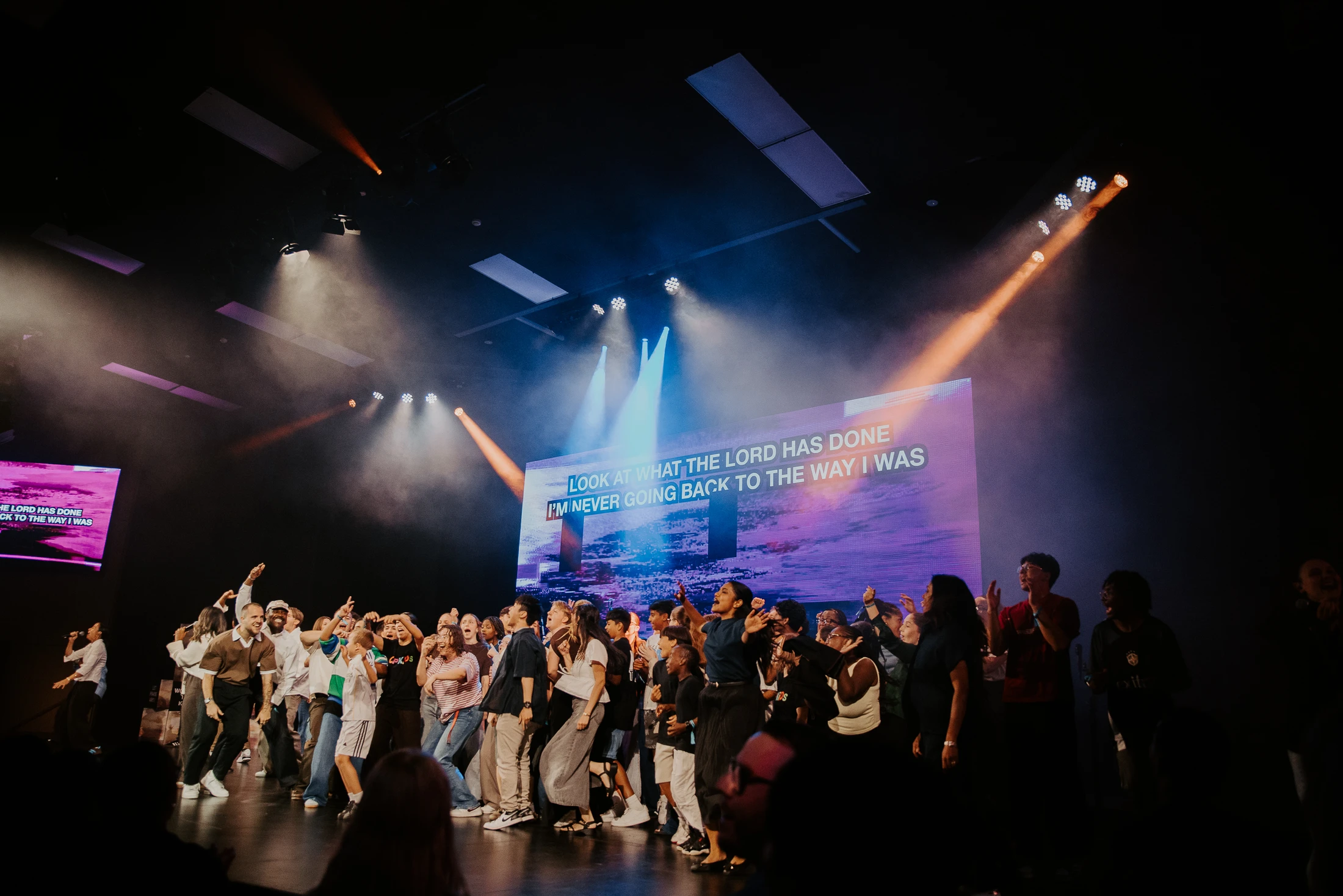 A diverse group of people sing and dance joyfully on a brightly lit stage. A large screen behind them displays worship lyrics under blue and orange spotlights.