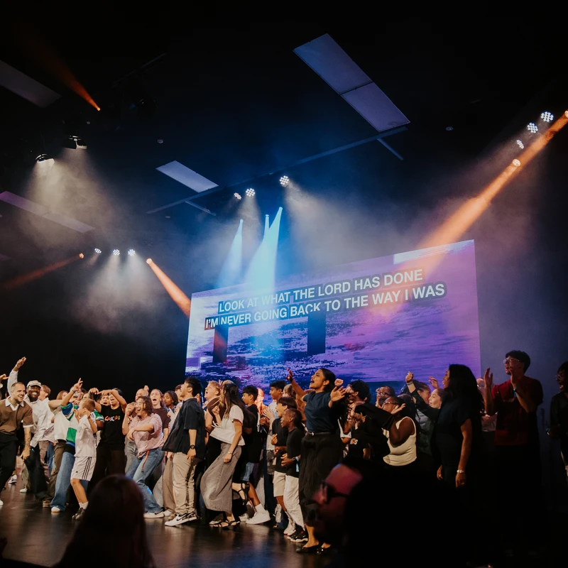 A diverse group of people sing and dance joyfully on a brightly lit stage. A large screen behind them displays worship lyrics under blue and orange spotlights.
