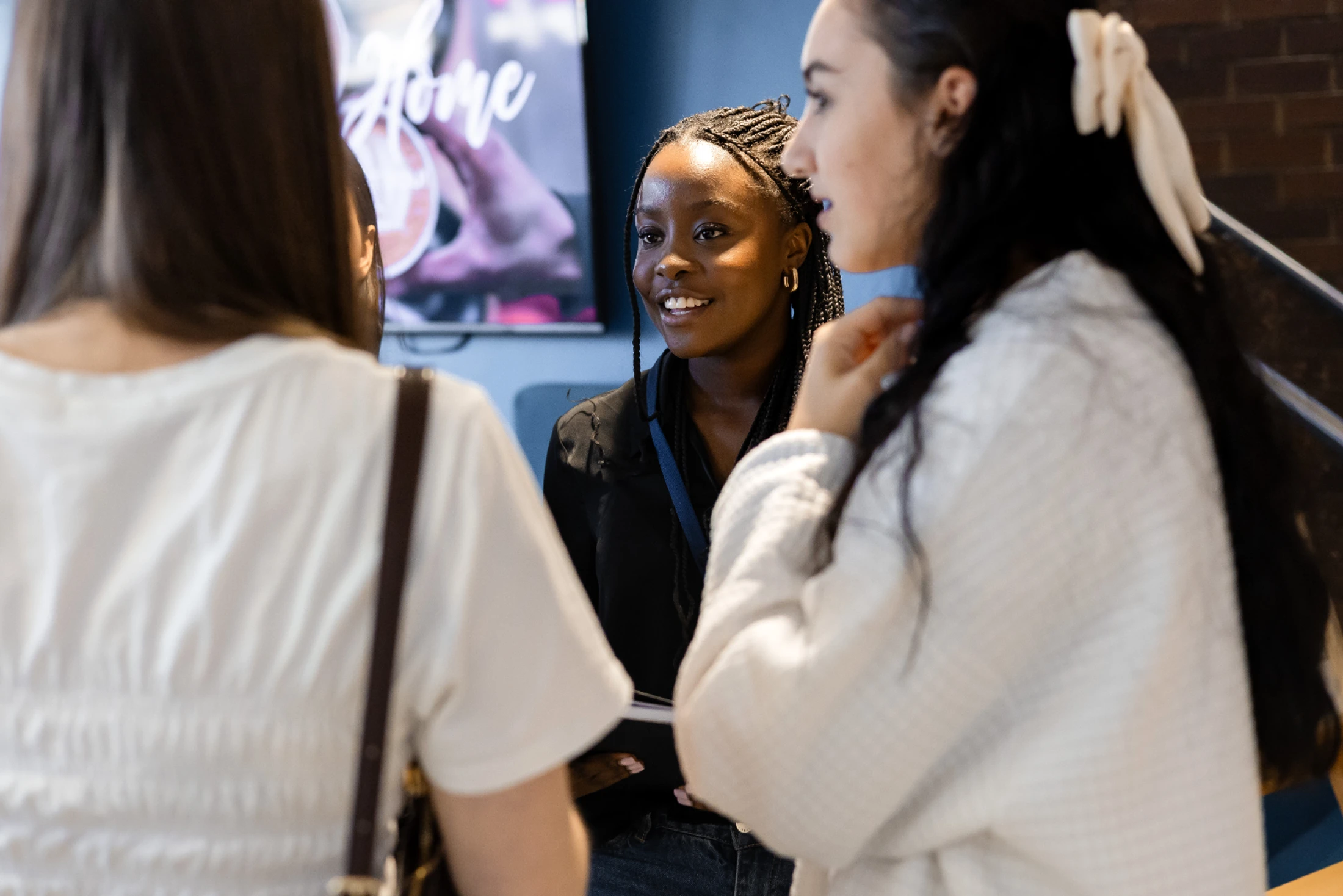 A young Black woman with braids smiles as she listens to two other women during a conversation in an indoor setting.