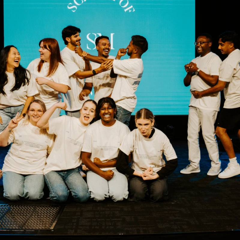 A diverse group of young adults in matching t-shirts laugh and pose for a photo on a stage in front of a large teal screen, radiating happiness.
