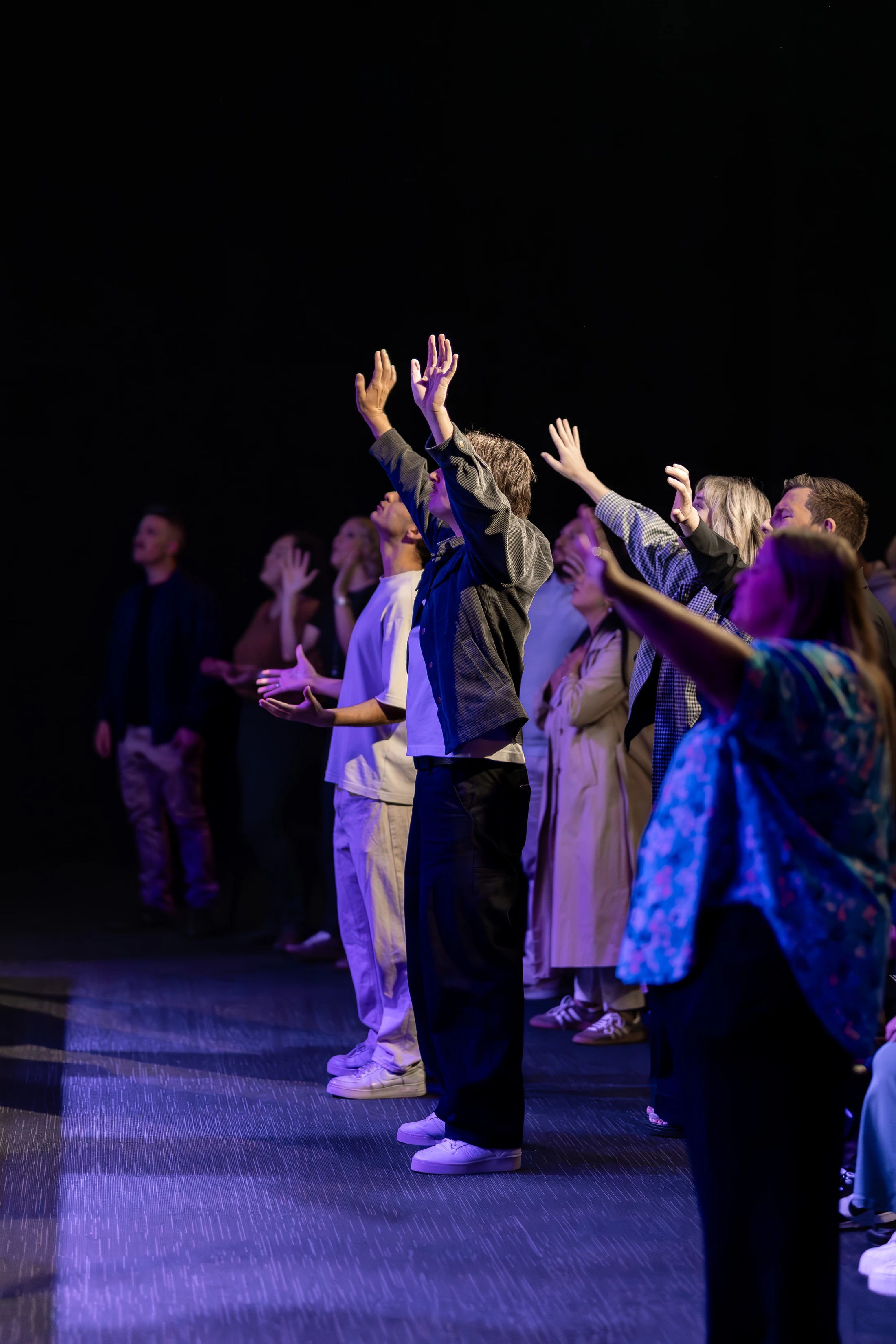 A side view of a group of people standing on a dimly lit stage with purple lighting. They are looking up with their hands raised in a gesture of worship.