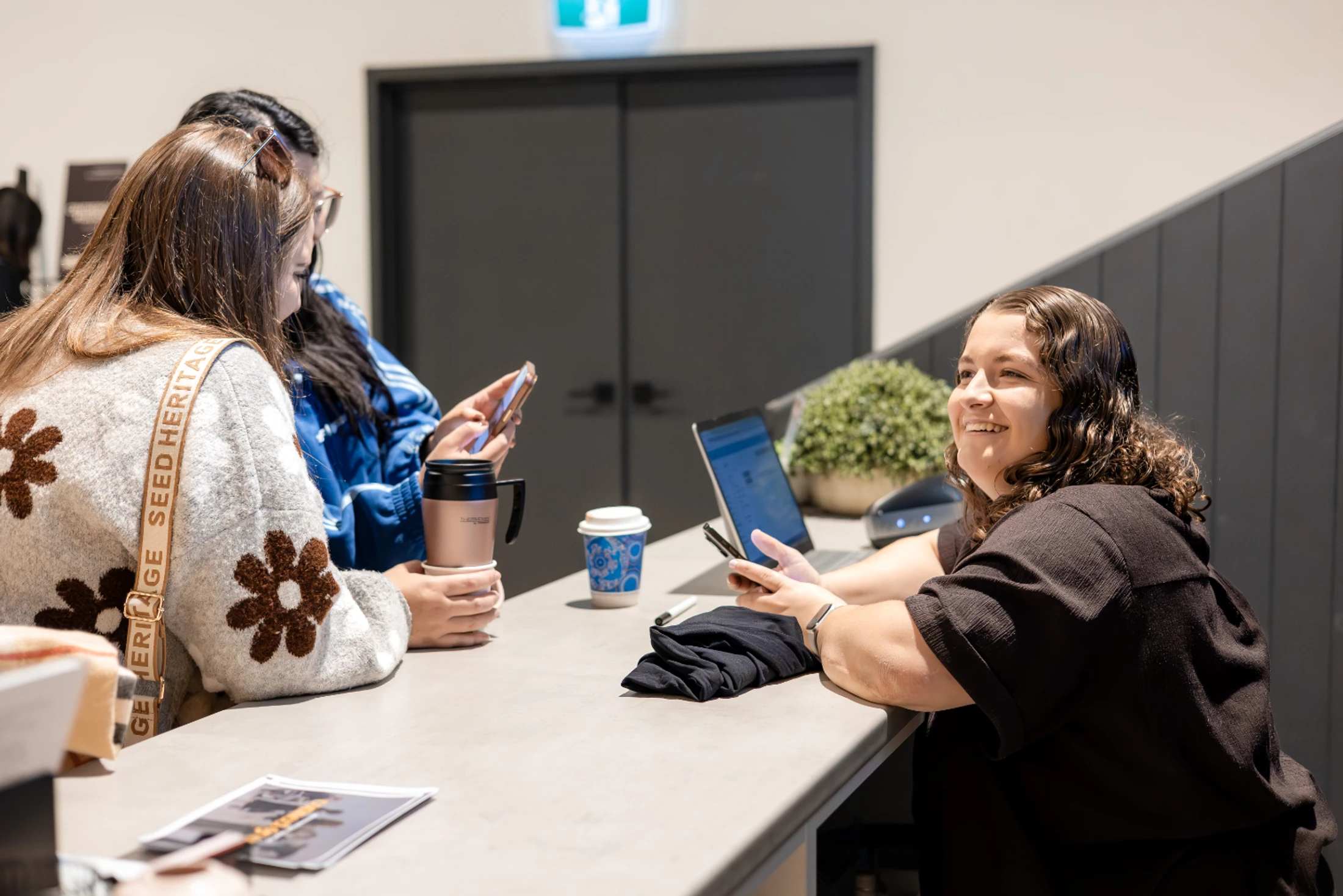 Three young women stand at a grey counter, engaging in conversation. One smiles warmly while the others look at their phones. Coffee cups are on the counter.