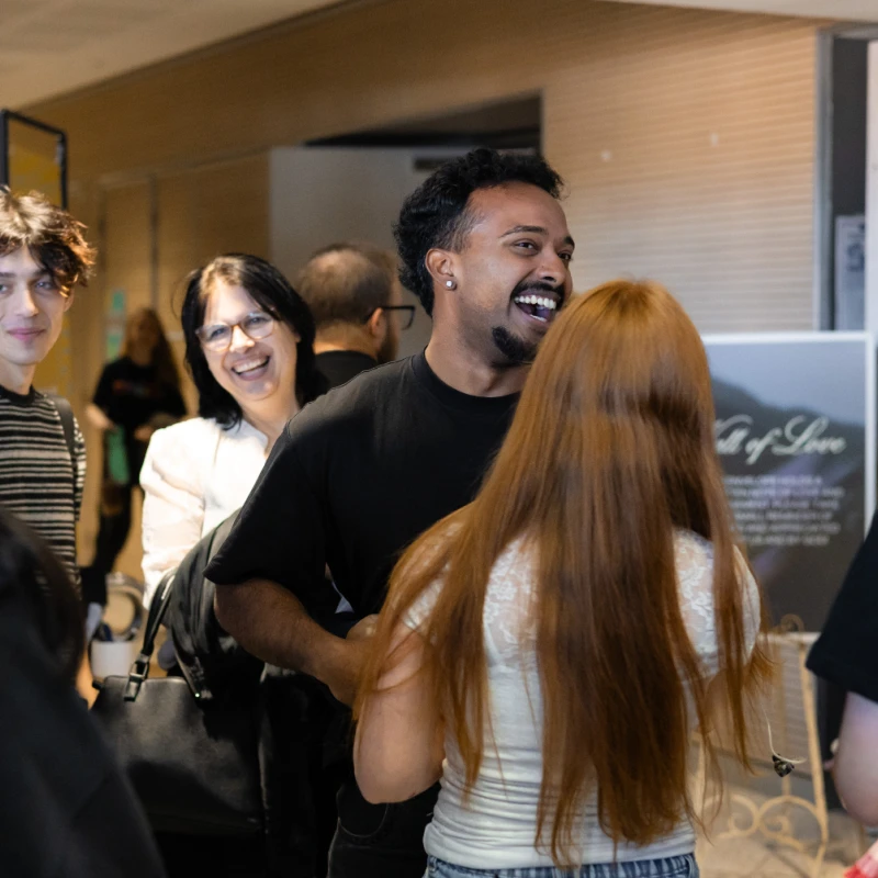 A diverse group of people talking and laughing together indoors. A man in a black shirt is the focus, smiling broadly while interacting with others.