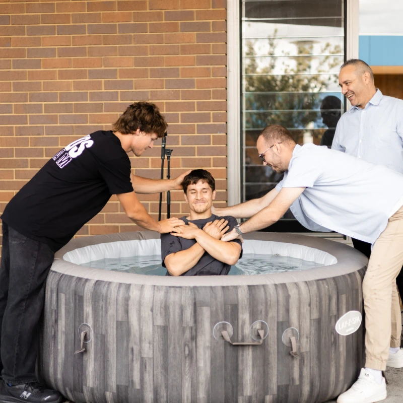 A young man sits peacefully in an inflatable pool with arms crossed as two men on either side prepare to baptize him in an outdoor ceremony.