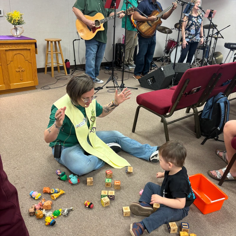 A woman in a stole sits on the floor with a toddler, playing with blocks and toys, while a band leads worship in the background of a church service.