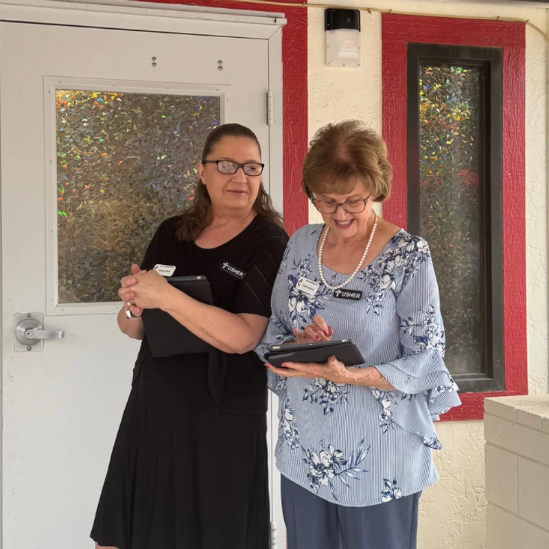Two women wearing usher name tags stand outside a church door. They are both holding tablets, and one woman is actively using hers.