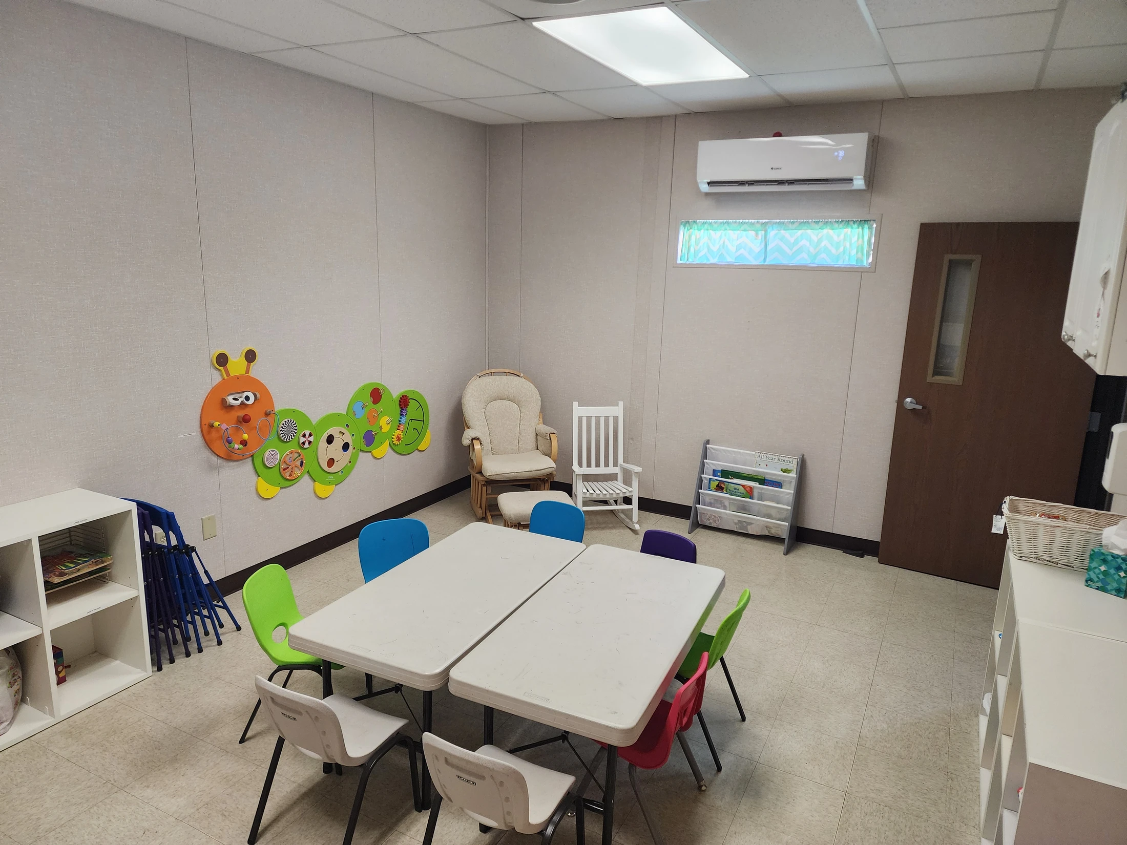 A brightly lit classroom for young children, with small colorful chairs around white tables and a caterpillar activity toy on the wall.