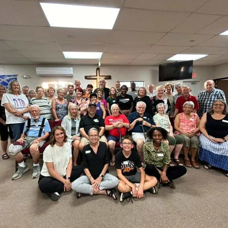 A large, diverse group of people of all ages posing for a photo inside a church hall. They are standing, sitting, and smiling at the camera.