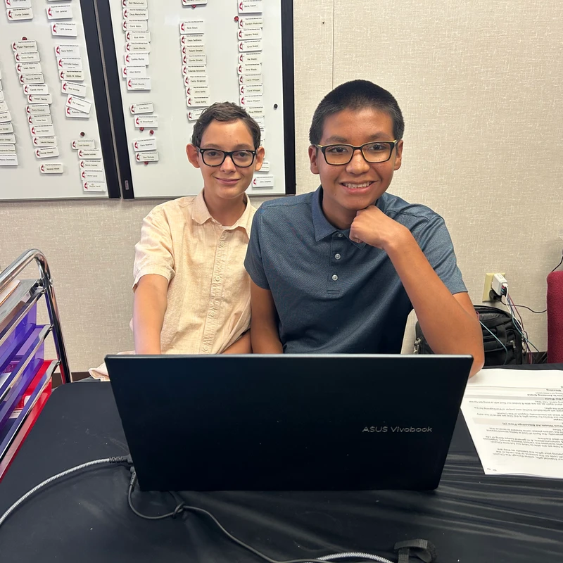Two smiling boys with glasses sit side-by-side at a desk with a laptop. In the background is a white board covered with magnetic name tags.