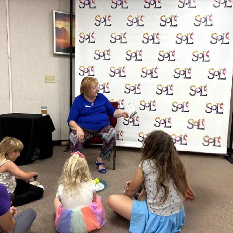 An older woman in a blue top sits in a chair and reads a book to three young girls sitting on the carpet in front of her during storytime.