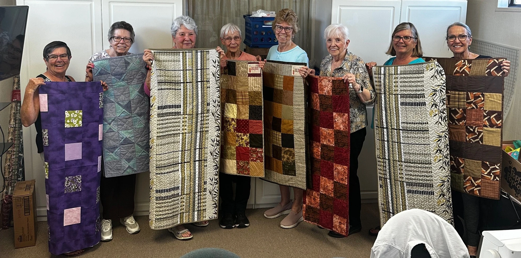 A group of eight smiling women stand indoors, each holding up a unique, handmade quilt they have created, showcasing various patterns and colors.