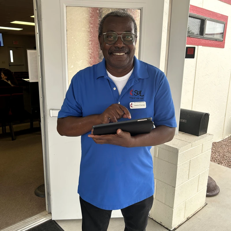 A smiling older Black man with glasses and a mustache, wearing a blue polo shirt, stands in a doorway holding a tablet.