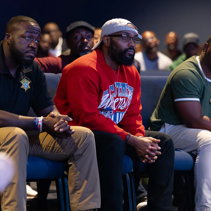 Three Black men sit in the front row of an audience, listening with serious, focused expressions. The man in the center wears a red sweatshirt and glasses.