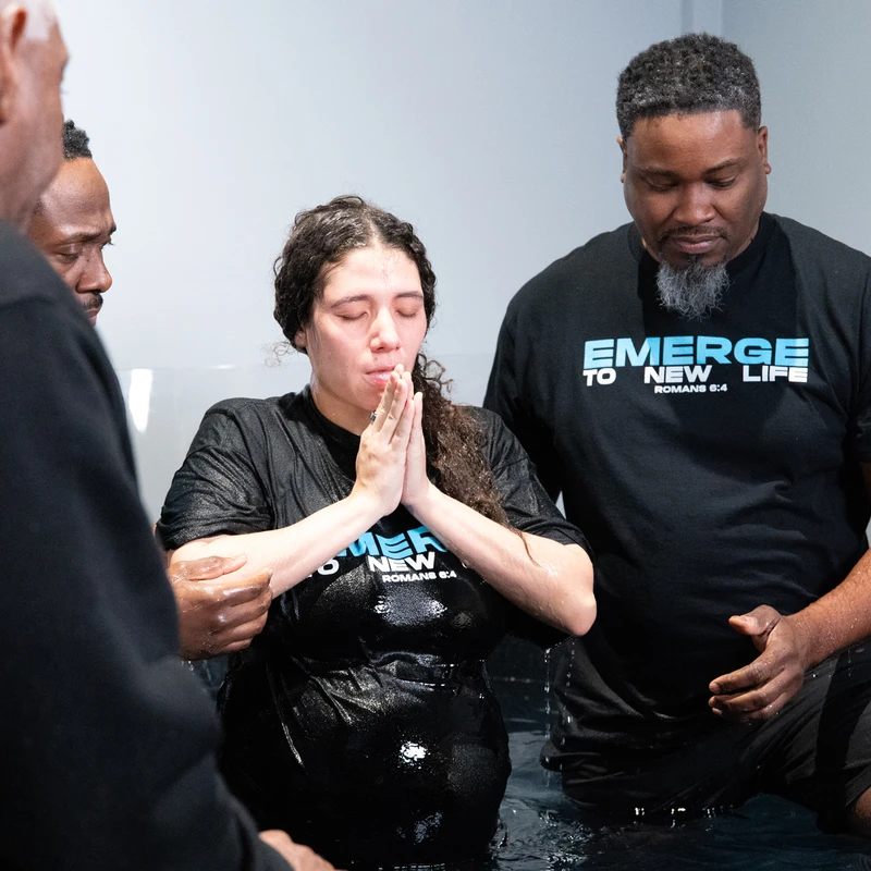 A woman with her eyes closed and hands in prayer stands in a baptismal pool, supported by two men during a Christian baptism ceremony.
