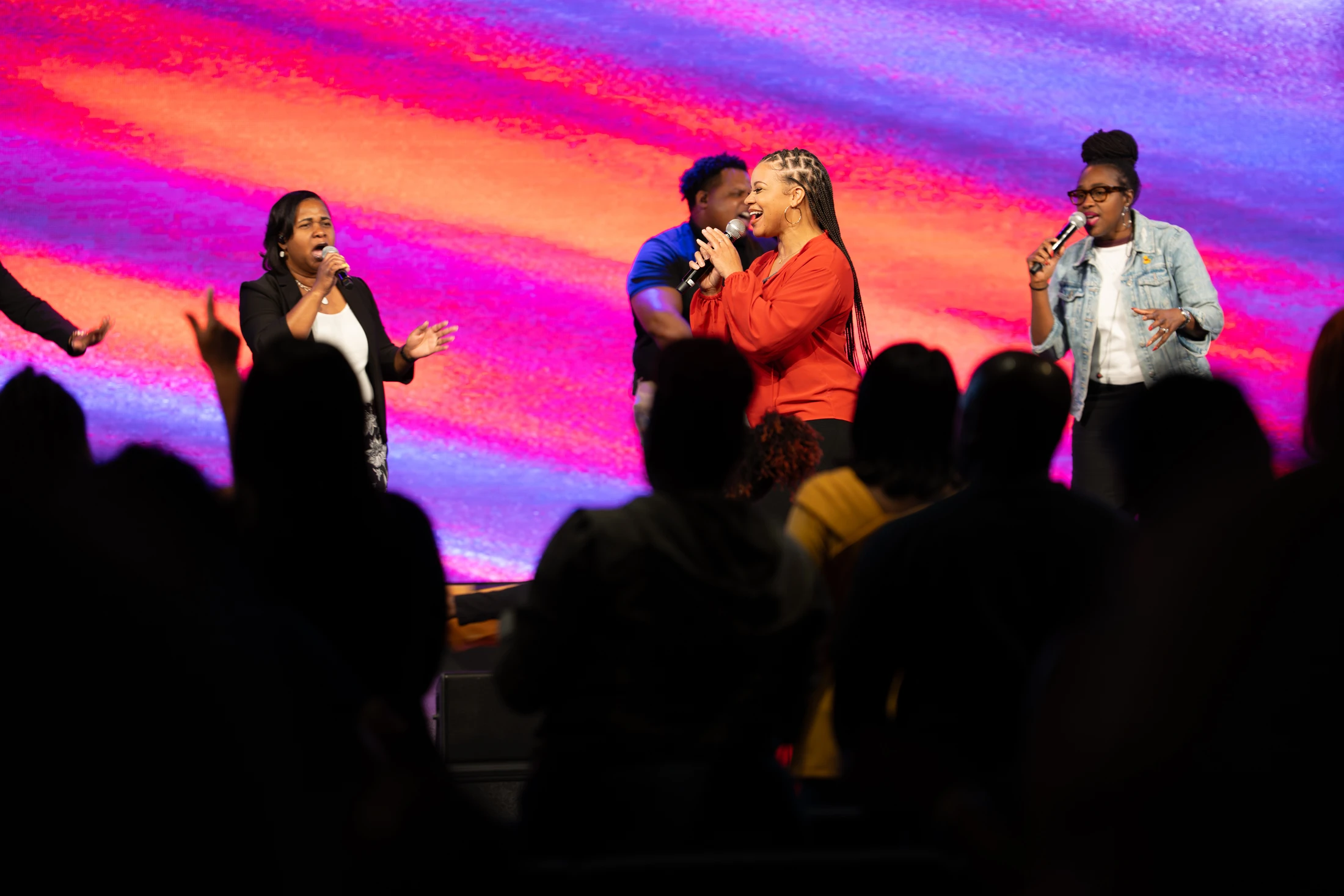 Several Black singers perform on stage with microphones in front of a colorful pink and purple backdrop, as a silhouetted audience watches from below.