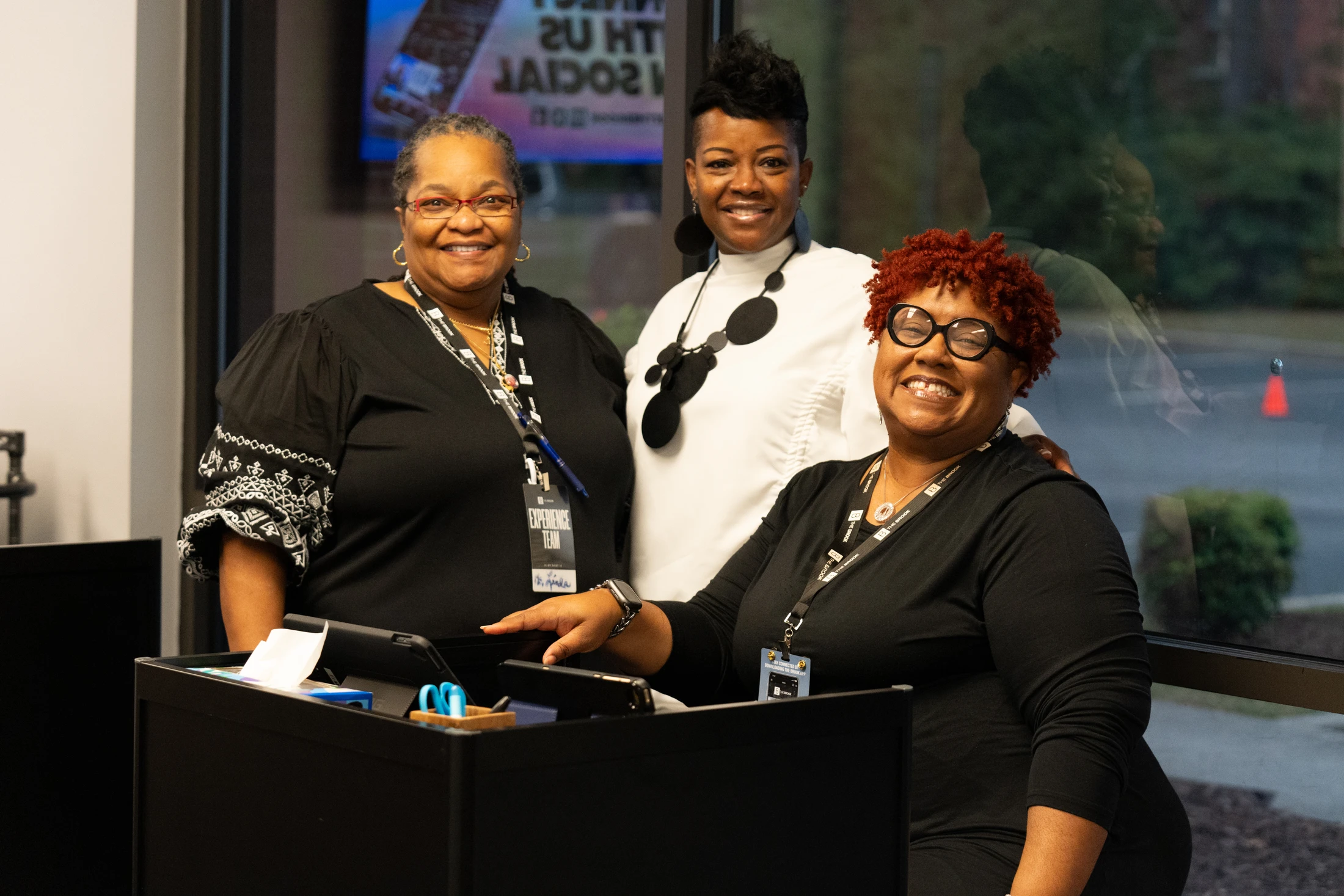 Three smiling Black women pose together for a photo indoors. Two are standing and one is at a lectern. All are wearing lanyards with event badges.