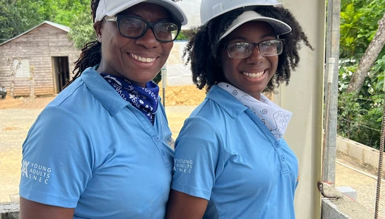 Two smiling Black women wearing matching blue shirts and white hard hats pose for a photo at an outdoor construction site while working on a cinder block wall.