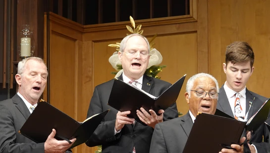 Four men of varying ages in black suits stand side-by-side, holding music binders and singing with open mouths in a wood-paneled room.