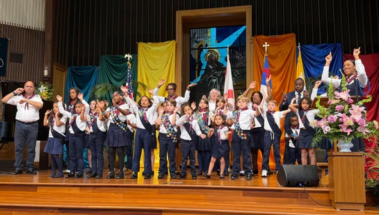 A large, diverse group of children and adults in scout-like uniforms perform on a wooden stage in a church, singing and using hand signs in unison.