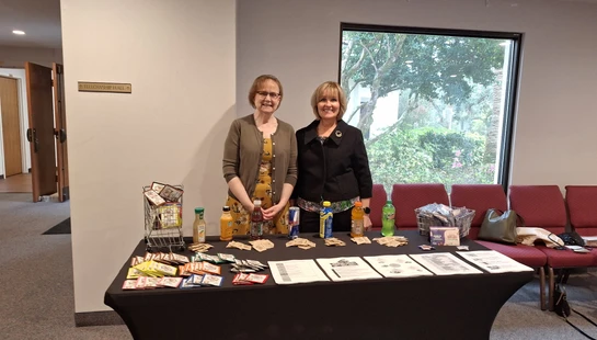 Two women smile behind a table covered in a black cloth, displaying various drinks, pamphlets, and informational papers in what appears to be a fellowship hall.