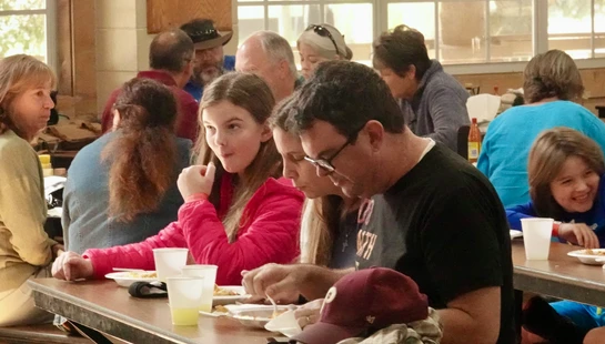 A family eats together at a long wooden table in a busy hall. A young girl in a pink jacket sits next to her father, who is wearing glasses.