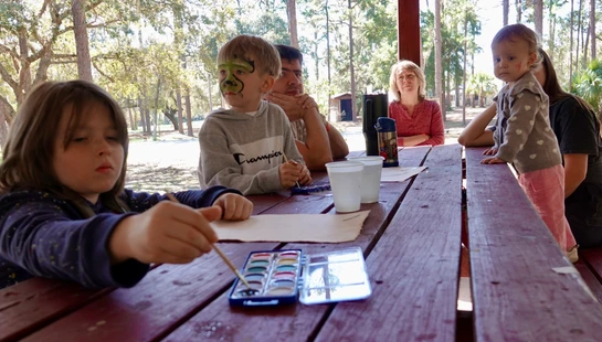 A family sits at a long red picnic table in a park. Two children are painting with watercolors, and one boy has green face paint on.