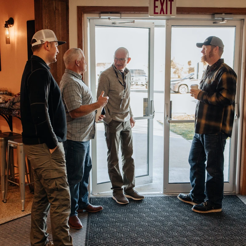 A group of four casually dressed men stand together in an open doorway, talking and smiling. The light from outside illuminates the entrance.