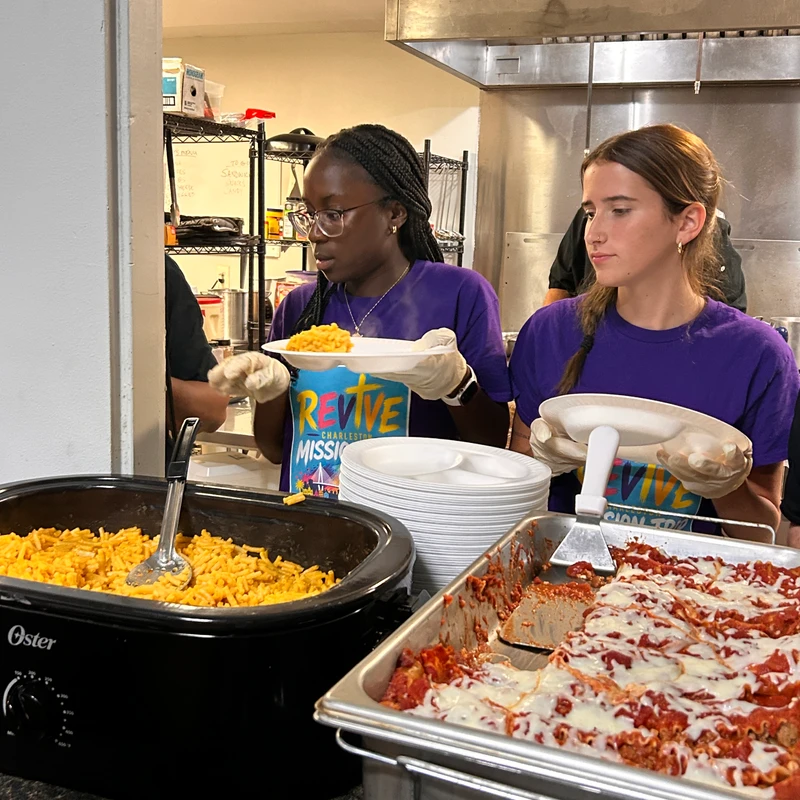 Two young women in purple t-shirts and disposable gloves serve macaroni and cheese and lasagna from warming trays in a kitchen setting.