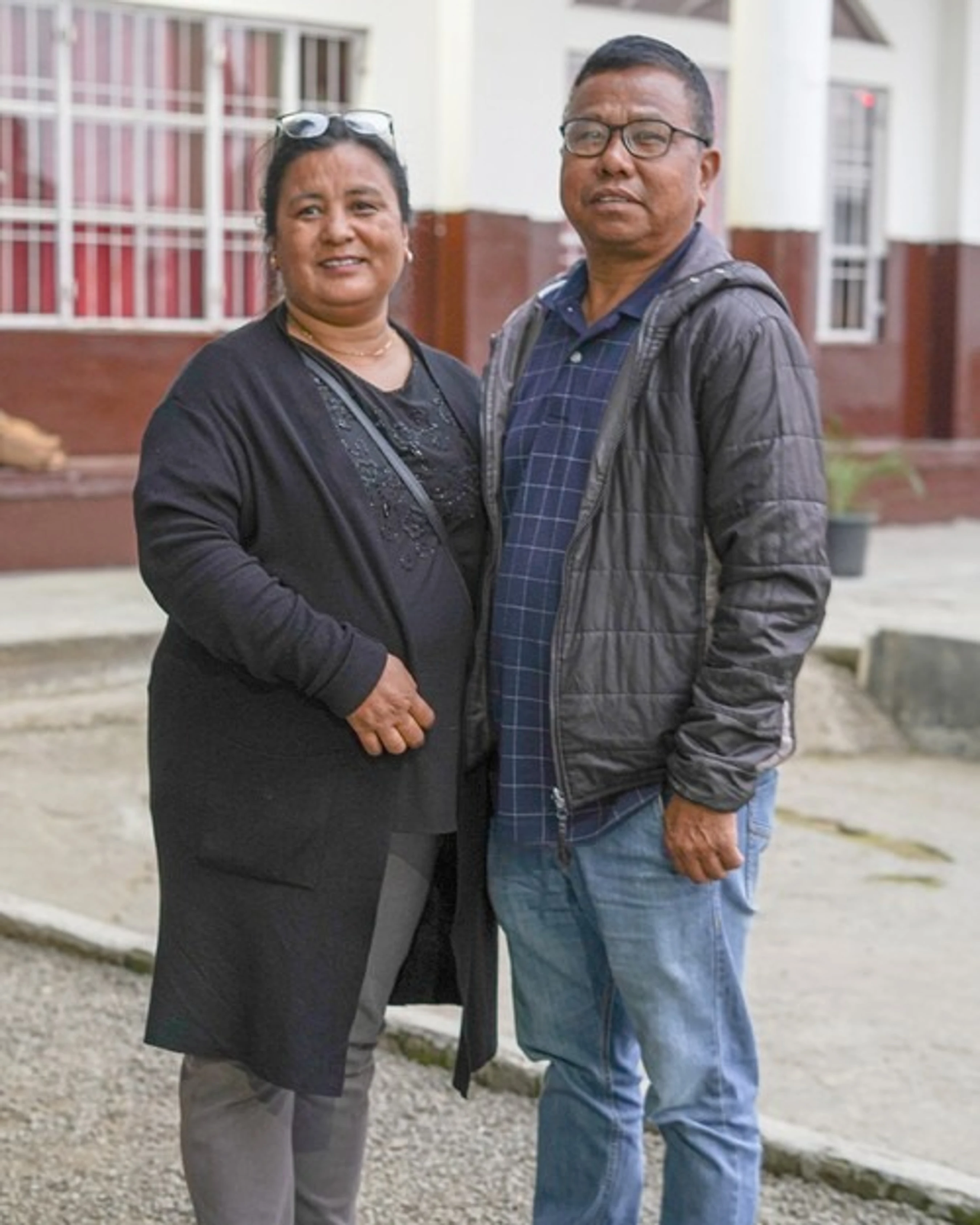 A middle-aged man and woman stand side-by-side and pose for a photo outdoors in front of a building. The woman is smiling at the camera.