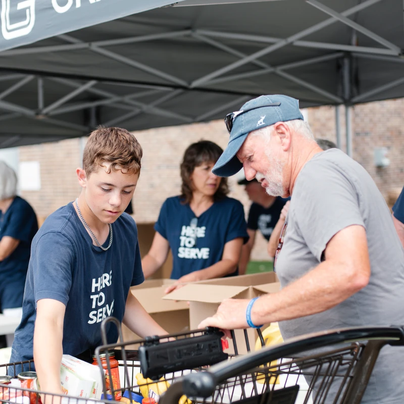 A young boy and an older man volunteer together, packing food from a shopping cart into boxes. Other volunteers work in the background under a tent.